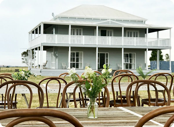 Photo shows brown bentwood chairs and a rustic trestle table, setup in front of a farm house ready for a backyard wedding